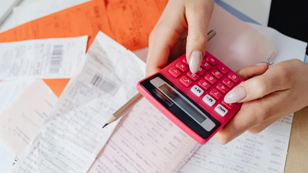 A woman's hands hold a red calculator over a pile of receipts and papers
